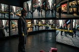 A young man, working as a janitor in a museum with wall-to-wall TV screens, stands there, looking at the same scene from different angles, viewpoints and sources, showing a crowded city street at a storefront. Part of the sidewalk is sectioned off with police tape, and inside that space is a raspberry milkshake,