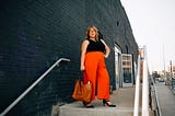 Allison stands on a cement staircase in an industrial neighborhood in downtown Denver. She is looking confident in high waisted wide-legged red pants and a blank tank top. She carries a brown leather bag, and an air of knowing her worth. Her face says quiet confidence.