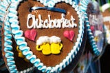 A large cookie decorated with the word “Oktoberfest,” featuring two red hearts and a pair of yellow beer mugs in the center, topped with bubbles.