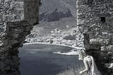 A young woman wearing a summer dress peers through a ruined stone arch at a coastal village far below.