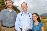 Paul Farmer stands between two GlobeMed students. He is wearing a long-sleeve, light blue button up shirt with a stethoscope slung around his neck. To the left is a student who is taller than Paul, has some scruffy facial hair, and is wearing a short-sleeve, gray button up. To the right is a student who is shorter than Paul and is wearing a blue patterned top with a square neckline. They are all smiling at the camera, with a rural landscape in the background.