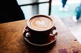 A cup of coffee on a worn out wooden table. The coffee has a heart drawn in milk, and is in a red mug on a matching saucer.