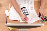 A picture taken from above of a desk on which a screen and paper written on in front of it, an iPhone and two hands can be seen.