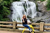 White woman with red hair sits on an yellow walking bike on a bridge in front of a water fall.
