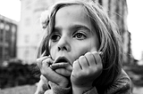 A black-and-white image of a light-skinned young girl. A close-up on her face set against a blurry background of old-brick apartment buildings. She has her head resting in her hands and she’s pulling her cheeks down, forming her face into a exagerrated frown.