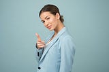 woman in blue coat, dark hair, pointing at camera, blue background