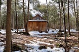 A cozy cabin in the woods, blanketed by snow, with a peaceful winter scene of trees and white ground surrounding it