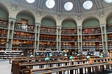 Vast BNF Richelieu library with vaulted ceiling, ornate arches and three levels of shelves overflowing with books. Reading tables with green lamps are neatly arranged, inviting study and reflection.