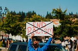 Protester holdiong adn “End Racism” sign at a rally. The author has used Microsoft Paint to create a big black “X” over the sigm.