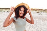 Smiling black woman on the beach, holding onto her hat.