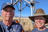 Two volunteers take a selfie in front of a metal and wood tower.