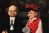 Two marionettes of an old couple — one man, one woman. Wrinkled from age, looking grouchy.