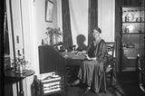 Black and white photograph of Eleanor Roosevelt seated at a wooden writing desk in a formal interior room. She is wearing a long satin dress and looking toward the camera while working at the desk, which holds papers and a small microphone. The room features tall curtains, framed artwork on the walls, a side table with flowers, and a wooden cabinet displaying plates and decorative items.