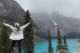 Woman standing on a rock facing mountains in the fog and a bright blue lake with arms spread wide open.