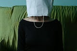 A woman sits on a green sofa, her head covered by a white paper bag. She’s wearing a black top and has her hands in her lap.