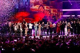 Wide, vibrant shot of a star-studded tribute concert: dozens of artists and celebrities line up on a brightly lit stage under purple and red spotlights as multicolored confetti rains down, performing in front of a packed arena crowd holding up smartphones.