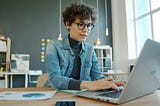 Small-business owner works at a laptop in an organized office with charts and notes visible, illustrating workflow management and business systems. Photo by Vitaly Gariev on Unsplash