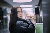 An attractive dark-haired girl in a black jacket is standing in an airport concourse, looking back over her shoulder towards the camera.