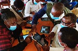 A group of adult learners holding tablets gather around a table to study