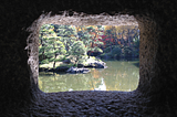 A photo taken through a Japanese stone lantern, looking to a bunch of small green trees on a grassy mound in the middle of a murky, green-brown lake. Additional trees fill the background. It’s a bright, sunny day. The image is framed by the darkness of inside the stone lantern.