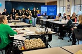 Six high high school students stand in front of a classroom giving a presentation to several adults sitting at desks.