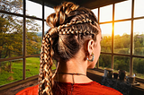 Side view of blonde, teenaged girl with elaborate French braids, looking out a window as the sun rises over a lush field in summer.