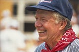 A closeup photo of former President Jimmy Carter, at a work site, wearing a neckerchief and baseball cap.