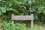 A wooden sign reads ‘Derwent Valley Heritage Way River Trent 55 miles’. Behind are green foilage and trees.