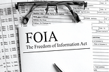 Papers on a desk with a pair of reading glasses with one of the papers reading “FOIA: The Freedom of Information Act”.