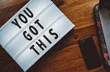 A lighted sign that says “You Got this” sits on a surface next to a smartphone.