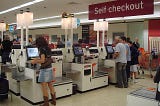 Three self-checkout machines at a store with a woman purchasing something at the first one and a man and son at the 3rd one