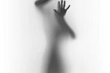 A photograph of an individual, with their hand pressed against the surface, is struggling to break free from the foggy white glass. Most of the photo is just a white background with just the trapped individual in the center. The individual’s face is unseen and the dark silhouette of the rest of their body can only be seen in the photo.