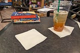 A small table in a Barnes and Noble Café. At the far left of the table, there are two books — and Ice Planet Barbarians book and Bluebeard’s Castle by Anna Biller. Plus two mystery digest magazines. In the foreground, there is a large cup of ice tea mixed with lemonade and some small napkins.