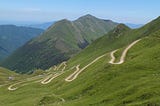 Green mountains under a blue sky, with a pale stone road zig-zagging up the mountain.