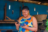 An African lady smiling while holding a phone in the right hand and a Point-of-Sale device in the left hand.