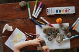 A kids hand on a desk splayed with sketch pens, paint, paper, rocks and crayon