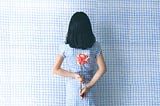 Back of a female-identified person holding a flower behind her back. She has shoulder length black hair. She is wearing a black and white dress, in front of a background that matches.