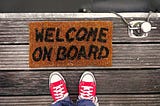 Foto de pés de uma pessoa com tênis vermelhos, visto de cima, no chão existe um tapete de boas vindas onde é possível ler “Welcome on board”