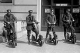 Four 20th Century postmen stand on their Autopeds, the electric scooters of their time.