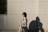 Photo of a woman walking in the sidewalk wearing a trench coat, sunglasses, coffee in hand, and looking cool!