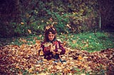 Young, brown haired woman with pony tail in a red sweater and fall colored scarf, sitting in a huge pile of Fall leaves, falling all around her in Sukhasana with prayer hands.