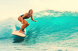 Blond female surfer riding a wave in a one piece swimsuit, crouched down, hand out to balance.