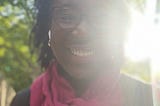 Selfie of the author, Scarlet Ibis James. She is backlit by brilliant sunshine and is wearing a red scarf. As usual, she has a dazzling smile.