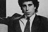 Jeffrey Epstein, wearing a dark suit and seated on a sofa with one arm resting on the top of the sofa, looks directly at the camera in a black and white photo