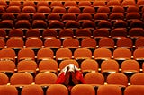 A single audience member sits in a big empty theater of orange seats. The audience member is hiding their head under their coat.