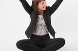 Woman in work attire — suit, and blouse — sits cross legged in front of a laptop and puts both arms in the air to celebrate