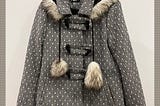 A black and white tweed jacket with a fur-trimmed hood, pom poms and toggles with the words, ‘a tweed jacket on a hook catches my eye’ and ‘“Try it on’, the story lady says, “Oh it suits you!”’ inserted in text boxes.