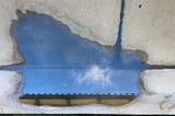 A puddle on concrete that reflects a wispy cloud in the deep blue sky, floating above roof tiles
