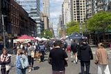 People walking on a pedestrianised avenue in New York City. It’s a beautiful day and the road is closed to cards because of a street fair. You can see the stalls on the street and many high rise buildings on either side of the road.