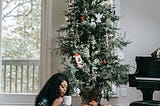 Black woman in pajamas enjoying drinking from a mug as she reads on the floor in front of her Christmas tree.